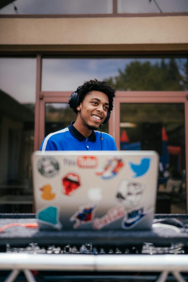 Young disc jockey in blue shirt smiling while mixing music on a laptop outdoors.