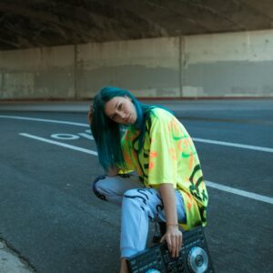 Young person with DJ equipment and vibrant outfit posing under a bridge.