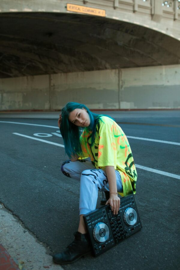 Young person with DJ equipment and vibrant outfit posing under a bridge.